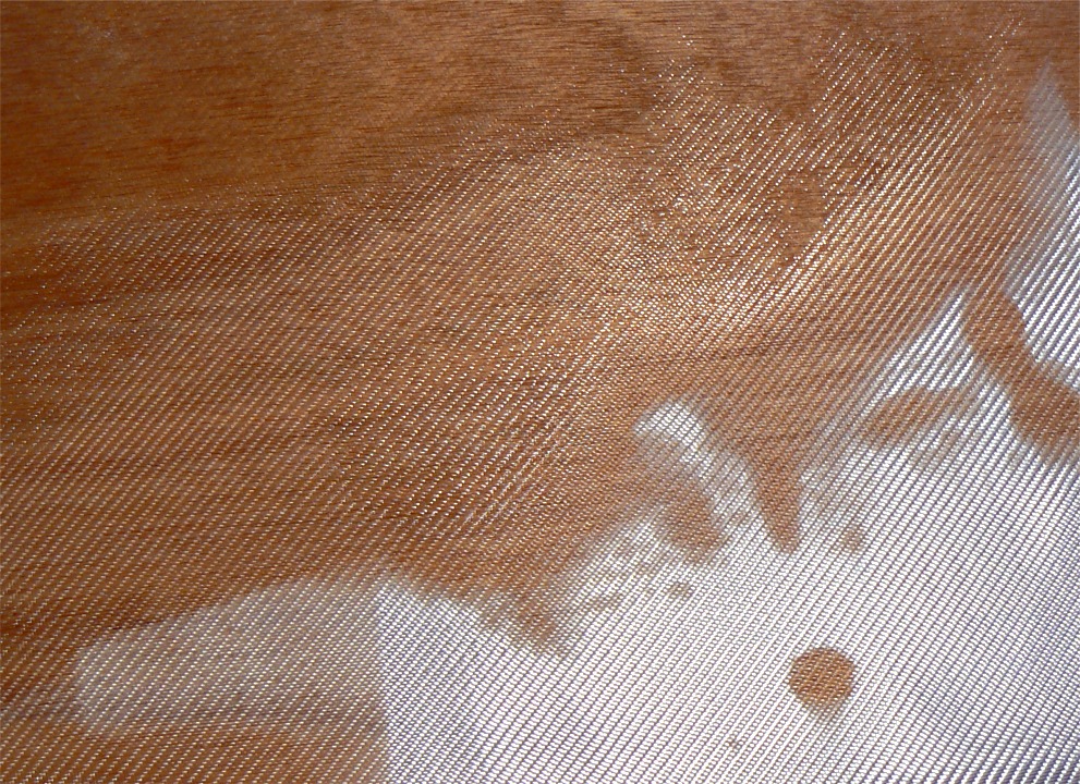 Gros plan sur le tissu de verre, à la limite des zones saturée et non saturée : le tissu devient totalement transparent lorsqu'il est saturé d'époxy, mais on doit voir sa trame très nettement. En haut à gauche, il y un peu trop d'époxy, on ne voit plus la trame du tissu. En bas à gauche, c'est l'inverse : le tissu est grisâtre, on ne voit pas le bois à travers. Il n'est pas totalement saturé, il faut lui apporter un peu plus d'époxy. Le reste du tissu en bas à droite est encore blanc car il n'a pas encore reçu d'époxy. 