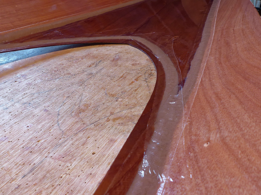 Cette vue de la surface intérieure du pont du Wood Duck Double montre le profil convexe des joints obtenu en les formant à l'aide d'une raclette en plastique souple. On voit aussi la bande de fibre de verre qui "arme" la surface de ces joints et étend leur "ancrage" sur le contreplaqué de part et d'autre du joint. Notez que le bois visible par l'ouverture du cockpit est celui de ma longue table de travail, sur laquelle j'ai percé beaucoup (mais alors beaucoup) de trous de sutures depuis 10 ans. Bonne nouvelle pour elle, les trous de la plupart de nos kits sont maintenant pré percés au moment de la découpe. 