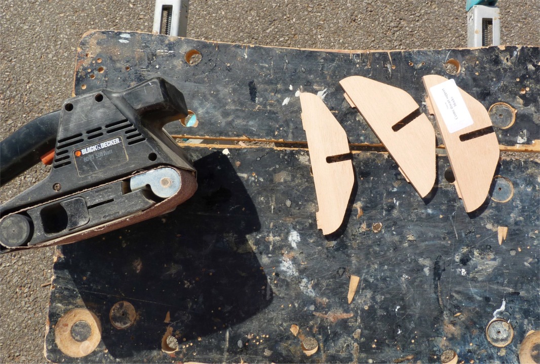 Voici les trois petits supports du banc central, qui s'encastrent dans les trois fentes du haut de la membrure centrale. Au préalable, je vais supprimer à la ponceuse à bande les ergots laissés par la machine de découpe numérique. Ces ergots servent à conserver une cohésion minimale des pièces dans la feuille de contreplaqué, afin de faciliter l'extraction des pièces après la découpe. 