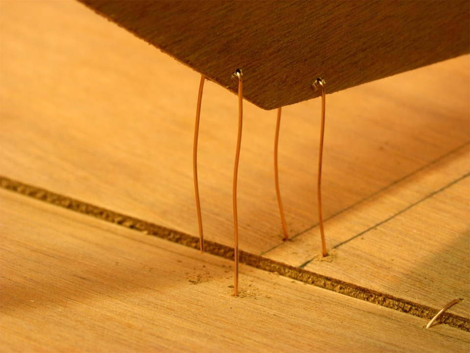 Gros plan sur les deux sutures de l'angle inférieur tribord de la cloison avant. J'ai inséré d'abord les sutures dans les trous percés dans la cloison, je les ai pliées en "U", et j'ai inséré chacune des pointes des "U" dans les deux trous percés à travers la sole. On voit d'ailleurs que l'extrémité la plus à gauche est plus courte n'est pas encore entrée dans son trou. Notez que l'on voit la tranche de la sole en surplomb par rapport au bordé. En effet, la sole est réalisée en contreplaqué de 9 mm d'épaisseur, tout comme les cloisons, alors que les bordés sont en 6 mm. On aperçoit une suture joignant le bordé à la sole au premier plan à droite.