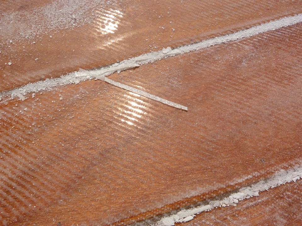 Gros plan sur la surface stratifiée après ponçage : la poudre blanche est surtout constituée d'époxy, mais il y a aussi un peu de fibre de verre, parfois en morceaux assez gros, comme la baguette que l'on voit au centre le l'image, qui fait 7 cm de long. Ce morceau provient de la lisière de la stratification, sur le joint entre les virures 2 et 3. Par endroits, le bord du tissu s'est légèrement relevé suite à la coupe, et j'élimine toute la fibre de verre qui n'est pas en contact avec le contreplaqué, car le composite est incomplet. 