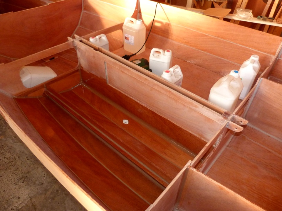Je viens de passer une dernière couche d'époxy claire sur toute la surface intérieure du réservoir de ballast tribord, ainsi que sur la face inférieure du panneau de fermeture et viens de le coller. J'utilise tous ces bidons pour le presser en place à cause du voilage résiduel qui gêne pour le coller uniformément. Au premier plan, je viens de passer la couche d'époxy claire sur le compartiment bâbord, au fond duquel on aperçoit le nable, collé juste avant. Je vais maintenant appliquer un filet d'époxy chargée sur tout le pourtour de contact du panneau avant de le mettre en place. 