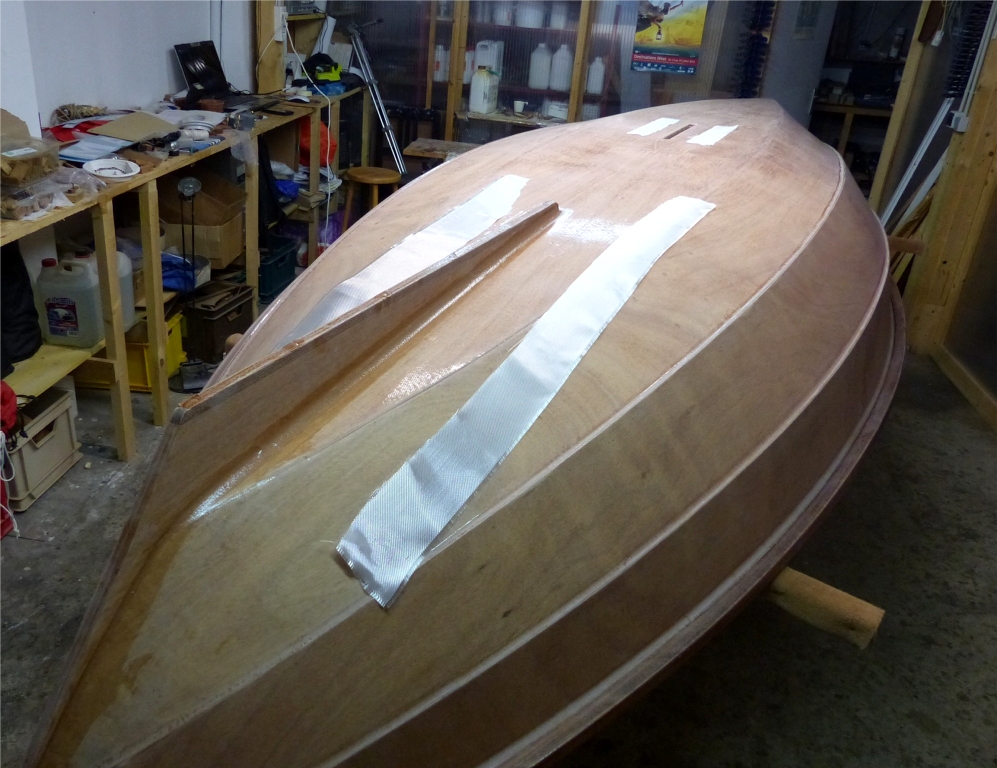 Je rentre le Skerry pour coller l'aileron, puis, après avoir avoir laissé un peu durcir l'époxy de ses joints-congés, je m'apprête à poser dessus les bandes de renfort que l'on voit ici, en même temps que de petites bandes sur le pourtour de l'ouverture du puits de dérive, qui se trouve actuellement à nu (ouh !) Toutes ces bandes sont découpées dans des chutes du tissu de verre de la stratification faite la veille. 