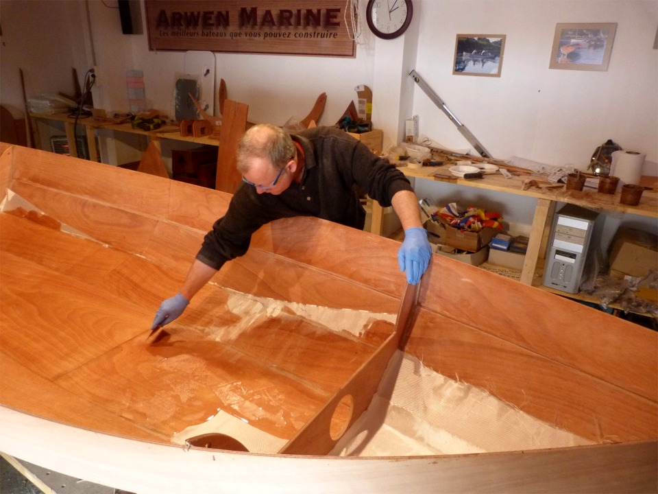 Hervé utilise la "squeegee" pour étaler la résine et la faire pénétrer dans le tissu de verre, qui devient ainsi transparent. On voit que la stratification ne couvre que la sole et la première paire de bordés. En effet, c'est le fond du bateau qui est à la fois le plus maltraité (à l'extérieur par les contacts avec des surfaces dures et à l'intérieur sous les semelles de l'équipage) et le plus solicité afin de rigidifier la coque. Cette stratification intérieure sera complétée par une stratification sur l'extérieur de la coque, également limitée à la sole et les deux bordés bas. 